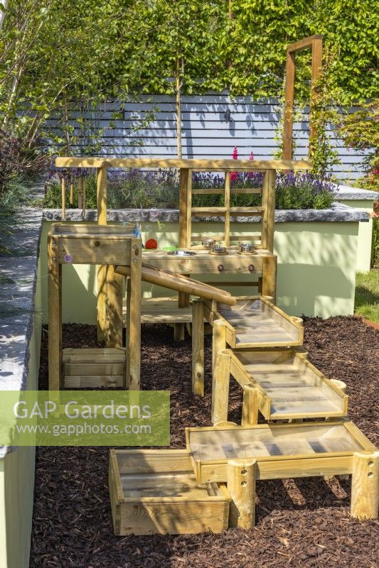 A small play corner in garden with a wooden mud pie kitchen and water run on floor of pine bark. June. Designer: Colm Carty, Bord Bia Bloom 2023