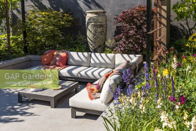 A sunken seating area with a comfortable corner sofa and low table surrounded by a perennial bed and two maple trees. A large ceramic vase decorates the background. Designer: Kevin Dennis
