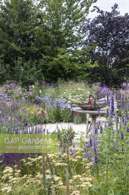 Perennial borders with salvia, achillea, agastache and Verbena bonariensis wrap around a wooden table with a trug full of gardening tools. Carol Klein, RHS Iconic Horticultural Hero - RHS Hampton Court Palace Garden Festival 2023