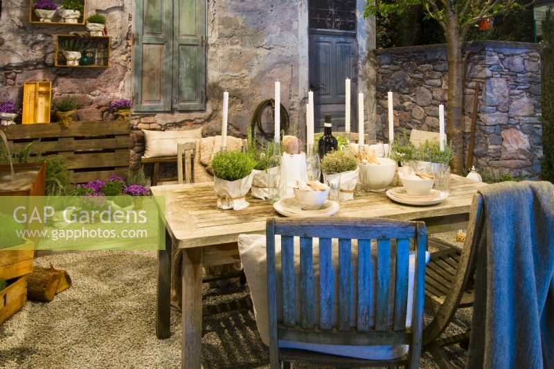 Decorative table prepared for a spring evening party with candles, glasses and herbs in pot in the contemporary Italian courtyard on gravel surface in front of the stone house. 
