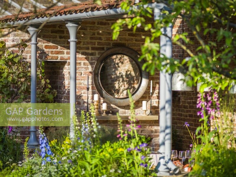 Circular mirror on brick wall  in an outdoor dining room. The Savills Garden, Designer: Mark Gregory, RHS Chelsea Flower Show 2023, May, Spring, Summer