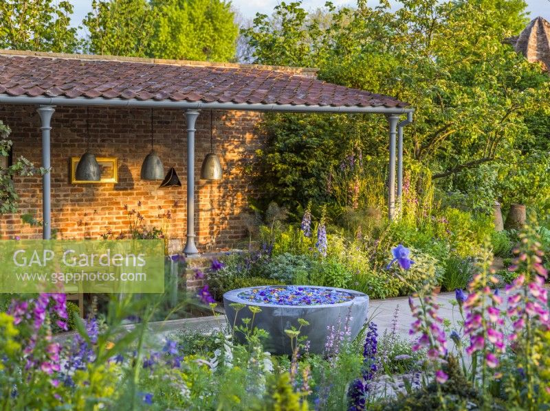 View across the garden with contemporary water bowl feature to sunken dining area. The Savills Garden, Designer: Mark Gregory, RHS Chelsea Flower Show 2023, May, Spring, Summer
