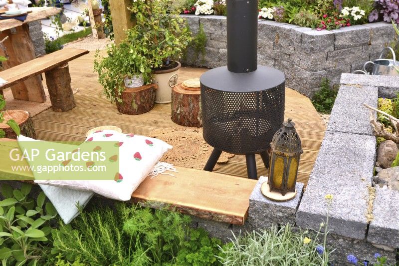 View of picnic area on wooden surface with modern chiminea behind raised bed made of  Connemara walling system in a woodland inspired garden. June
Designer: Mary Anne Farenden. Bord Bia Bloom, Super Garden, Dublin, Ireland.