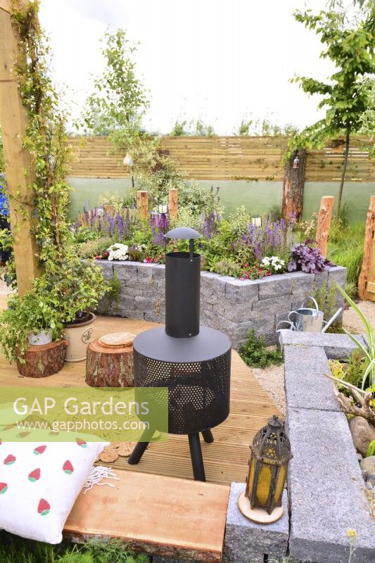 View picnic area on wooden deck with modern chiminea to a raised bed made of  Connemara walling system. Planted with perennials in a woodland inspired garden surrounded by a wooden planks fence. June
Designer: Mary Anne Farenden. Bord Bia Bloom, Super Garden, Dublin, Ireland.