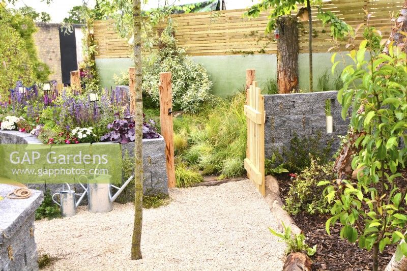 View of a open gate leads into a grass area for dogs with raised bed  made of Connemara wall system and gravel surfaces  in woodland inspired garden surrounded by a wooden planks fence. June

 


