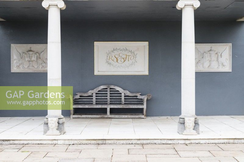 The interior of the Colonnade Court with Lutyens style wooden bench and plaques on the grey painted walls. The central plaque has initials of Roy Strong and Julia Trevelyan Oman. April. Spring. 