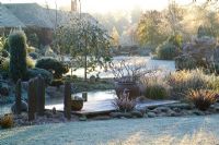 Early sunlight over the frozen pond and deck on a frosty winter's morning. Slate posts and a container of Prunus incisa 'Kojo-no-mai'. 