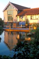 View across swimming pond to timber decking, outside eating area with table and chairs and converted barn with warm autumn sunlight reflected on building