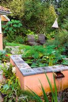 Raised terracotta painted pool water feature with cascade on terrace of exotic style garden with seating and tropical plants