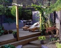 Garden with decking, Acer and Hosta in pots, loungers, pergola with vine, bamboo and raised border with blue grasses