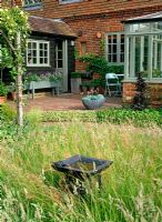 Meadow in small country garden with stone birdbath, brick terrace with container plants including Echeveria in terrazzo pot