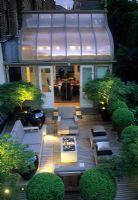 Overview of modern roof garden at night with lighting, table and chairs inside glass topped room and another outside. Containers of Buxus topiary spheres and Acers - Suleyman Garden, London 