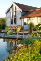 View across swimming pond to timber decking, outside eating area with table and chairs and converted barn with warm summer sunlight reflected on building - Carpe Diem, Bressingham, Norfolk