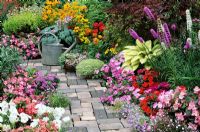 Bright and breezy summer bedding plants flank a cottage garden style path with an old watering can as a focal point. Petunias, Ageratum, busy Lizzies, Rudbeckias, fibrous rooted Begonias and Lobelia carpet around Hostas and Liatris 