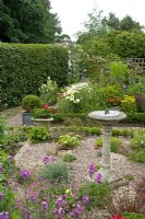 Mixed summer border graveled area with stone sun dial in centre shaped box in square container stone pot mature hedge and trees - Flower Garden at Coley Cottage, Staffordshire