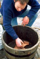 Man putting broken terracotta shards in bottom of wooden barrel to aid drainage, before planting it up