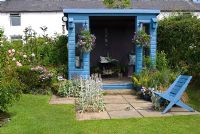 Blue painted wooden summerhouse with hanging baskets, and pots with blue seat on patio in cottage garden at New Row Cottages, NGS garden, Lancashire