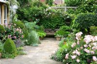 Terrace with wooden bench seat and decorative paving. Rosa 'Ispahan' to left and R. 'Felicia' to right. High trellis boundary with climbers.