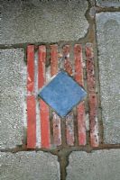 Marijke's garden. Decorative insert of a blue tile and red tiles on edge in a slabbed patio