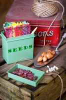 Fairtrade seed tin with seed packets, garden string and Runner bean seed 'Prizewinner stringless'. Onion sets, French Bean seed, stainless steel trowel on an upturned wooden crate