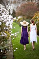 Two girls wearing summer dresses walking along a grass path between borders of shrubs and Magnolia