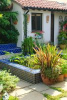 Paved courtyard with raised pool and seat clad in blue glazed tiles. Phormium in container.