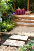 Gravel path and steps leading to oak deck with cushions, surrounded by lush planting. Small pond in foreground. 'The Yoga Garden' - Bronze Medal Winner - RHS Hampton Court Flower Show 2010
 