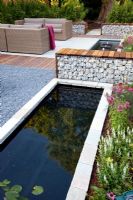 Small raised, rectangular pond and bench made from gabions in modern garden. Patio with sofas beyond.