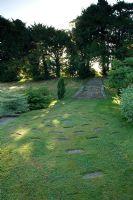 Round lawn with circle of paving slabs and steps up to mature trees. Robinson garden, Ousden House, Suffolk