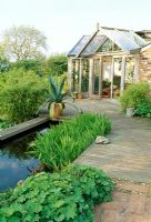 Timber and brick decking, pond and conservatory. Agave in container. Late Spring, Fovant Hut Garden, Wilts