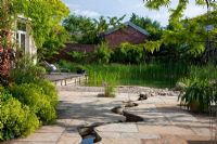 Natural swimming pool with pebble beach zone and a meandering canal leads through a flagstone paving towards the water. Planting includes Agapanthus, Alchemilla mollis, Allium christophii and Vitis 