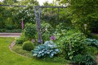 Mixed border in a shady part of a garden with carved sculpture. Planting includes Rosa 'Apfelblüte', Hosta 'Halcyon', Allium christophii, Buxus, Digitalis purpurea, Rodgersia and Rosa glauca 