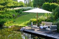 Relaxing area with parasol on wooden deck above a pond with primroses, flowering Aconogetum and a hedge encircle a lawn 