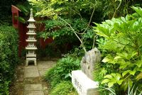Chinese granite pagoda in the Red Wall garden planted around with palm Trachycarpus wagnerianus and Edgeworthia chrysantha. 'Holding hands stone' on plinth in foreground surrounded by Bamboo, Fatsia japonica and Cladrastis sinensis. Beggars Knoll, Newtown, Westbury, Wiltshire, UK