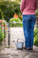 Woman wearing jeans and gardening boots standing at a garden gate with a vintage galvanised watering can