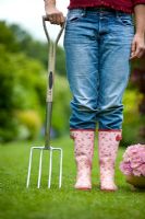 Woman wearing blue jeans and wellies holding a garden fork beside a wooden trug of pink chrysanthemum 