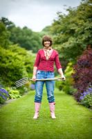 Woman wearing blue jeans and wellies standing on a lawned path carrying a garden fork 