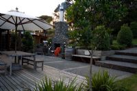 Deck and patio area with exterior fireplace. Corynocarpus laevigatus - Karaka, Pittosporum 'Mountain Green' - New Zealand