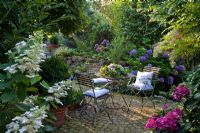 Wooden iron garden furniture with blue cushions on round paved rest area with shade loving plants, Buxus, Hydrangea macrophylla and Hydrangea paniculata
