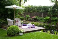White wicker garden furniture, a blanket with cushions and a parasol on a wooden deck next to a garden pond. Willow trellis and hedge in the background. Plants are clipped Buxus, Fern and Hydrangea