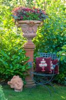 Terracotta urn on a plinth next to metal chair with cushions and terracotta head. Plants are Cotinus coggygria, Heuchera micrantha 'Palace Purple', Pelargonium and Prunus laurocerasus 