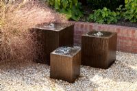 Gravel garden with wooden cube water features in gravel next to low brick wall. Epimedium x versicolor 'Sulphureum', Anemanthele lessoniana