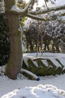 Snow covered garden. Children's play area with swing hanging from the branch of a Pinus - Pine tree and a tractor tyre used as a sandpit. Gowan Cottage in December