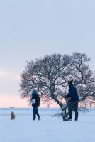 Father throwing snowballs at a boy, walking in the snow covered fields with a tree in silhouette against the sky in Westley Waterless. 
