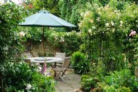 View of secluded corner of a formal town garden with circular patio table, parasol and chairs. Rosa 'Goldfinch'  growing on arch over path - Rhadegund House, New Square, Cambridge.