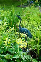 Heron statue beside pond surrounded by Primula grandiflora, Irises, Alchemilla mollis and ferns - Mindrum, nr Cornhill on Tweed, Northumberland, UK