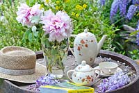 Vase of Paeonia and Deutzia with tea set, book and sun hat, Wisteria in background 
