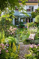 Stepping stones lead from the house to a round paved rest area with metal chair in a small garden with Rose 'Ballerina', Digitalis purpurea, Lobelia erinus, Lysimachia punctata, Nepeta Faassenii-Sorte and Petunia Surfinia