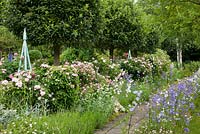 A paved path lined with wildflowers, roses, standards and plant supports - Rosa 'Duchesse de Montebello', Artemisia, Betula, Campanula persicifolia, Centranthus ruber 'Albus', Crataegus, Geranium sanguineum and Lavandula - Germany 
