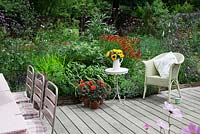 Lloyd Loom chair with cushion, small table with vase of Sunflowers in decked garden with densly planted border. Pots of Pelargoniums. Verbena bonariensis, Heleniums, Achillea, Nepeta, Rose bush with hips and Cardoon.
